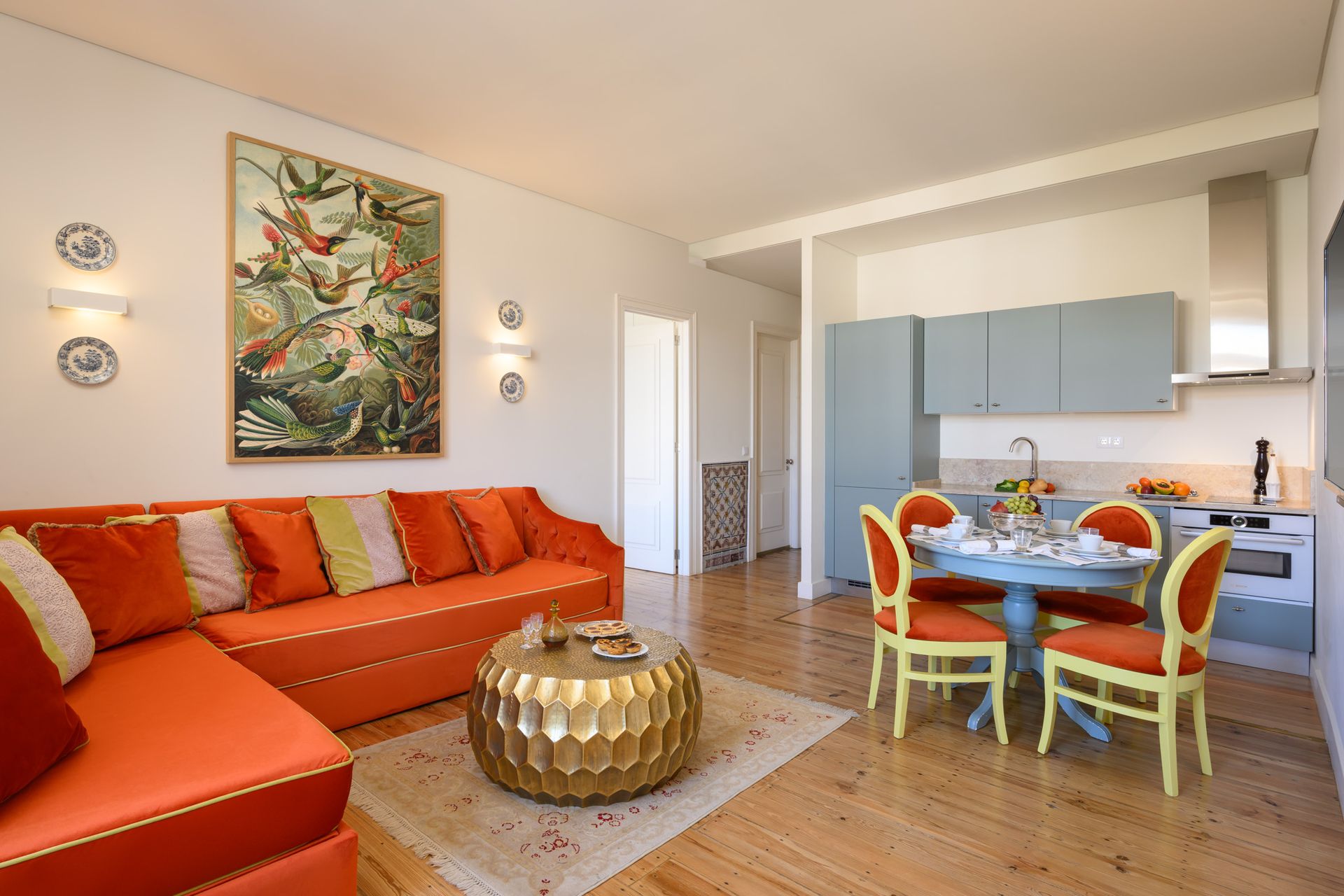 Bright orange living room with a sectional sofa, round dining table, and small kitchen.