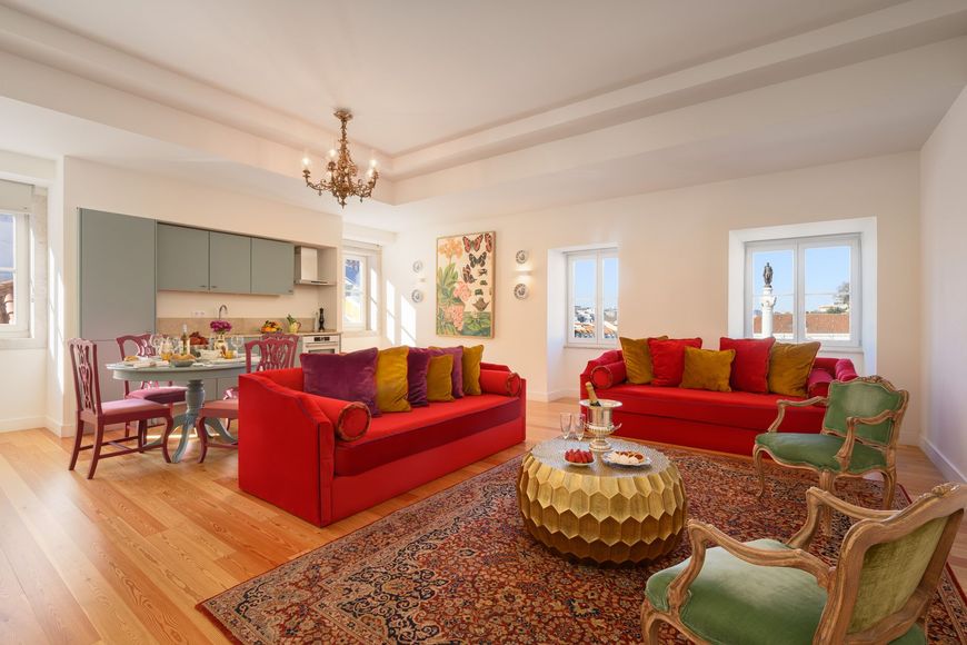 A bright, modern living room featuring a red sofa, a gold coffee table, patterned area rug, and an integrated kitchen.