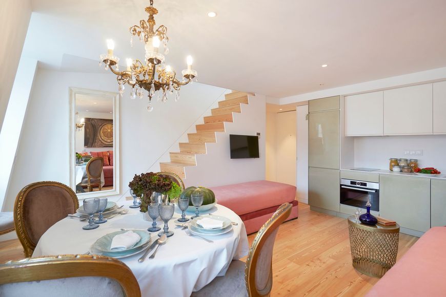 Small apartment with dining area, kitchen, and stairs to a loft; round table, chandelier, pale pink accents.