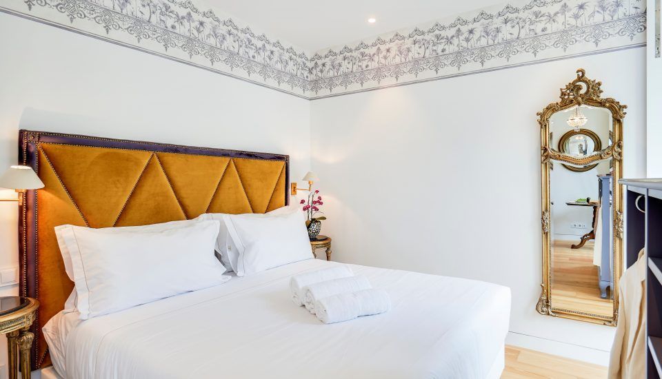 Bedroom with gold-toned headboard, full-length ornate mirror, white linens, and floral wallpaper border.