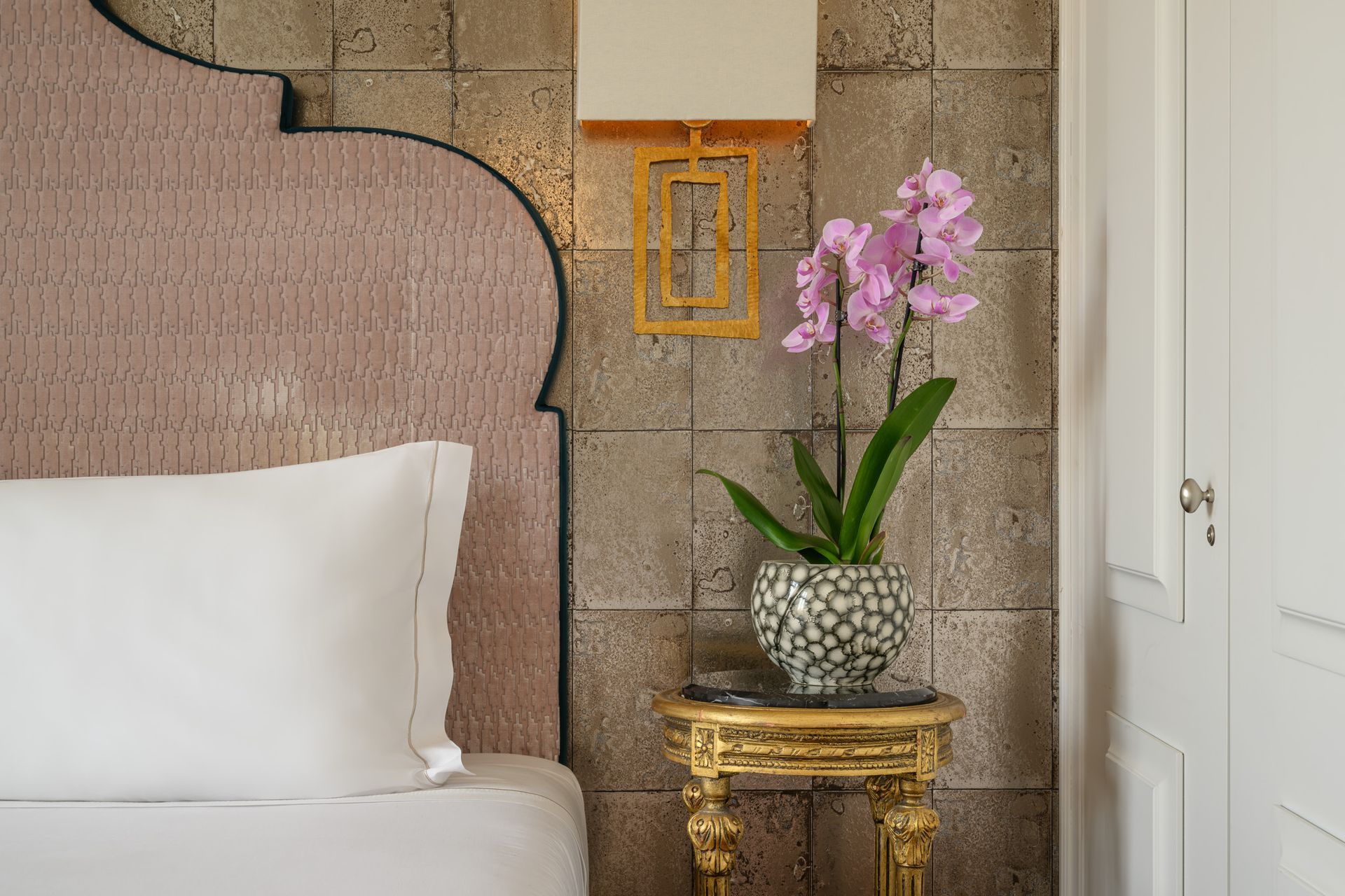 Bedroom with pink headboard, gold side table holding an orchid, lamp on textured wall.