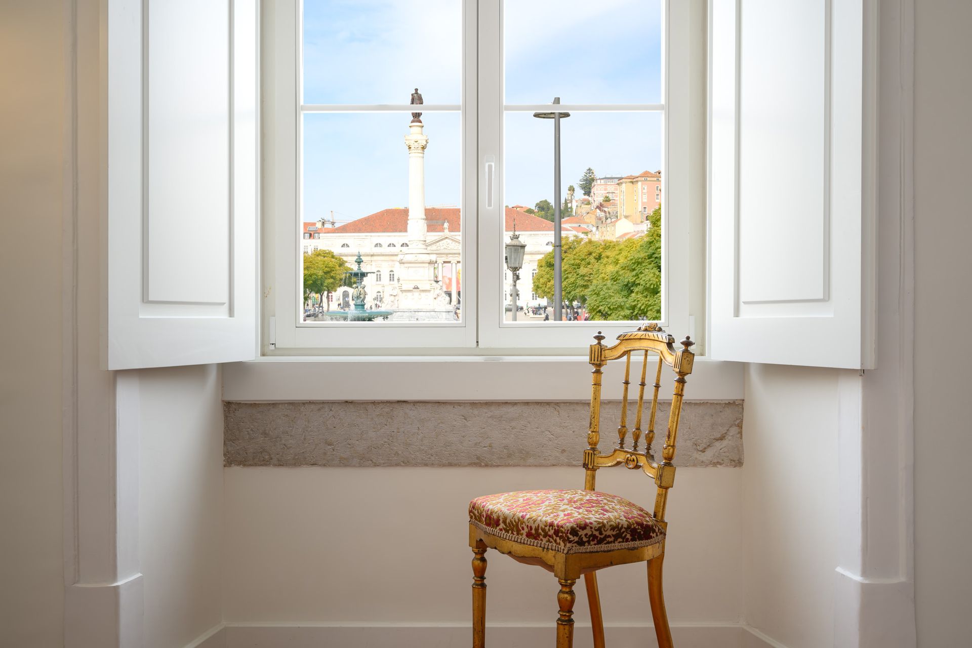 Golden chair sits before a window overlooking a plaza with a monument and trees.