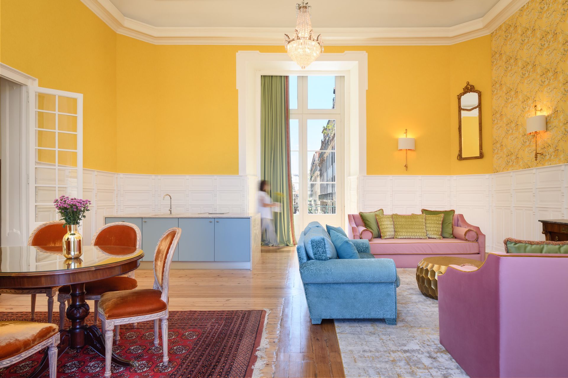 Living room with yellow walls, colorful furniture, and open doors with a person in the doorway.