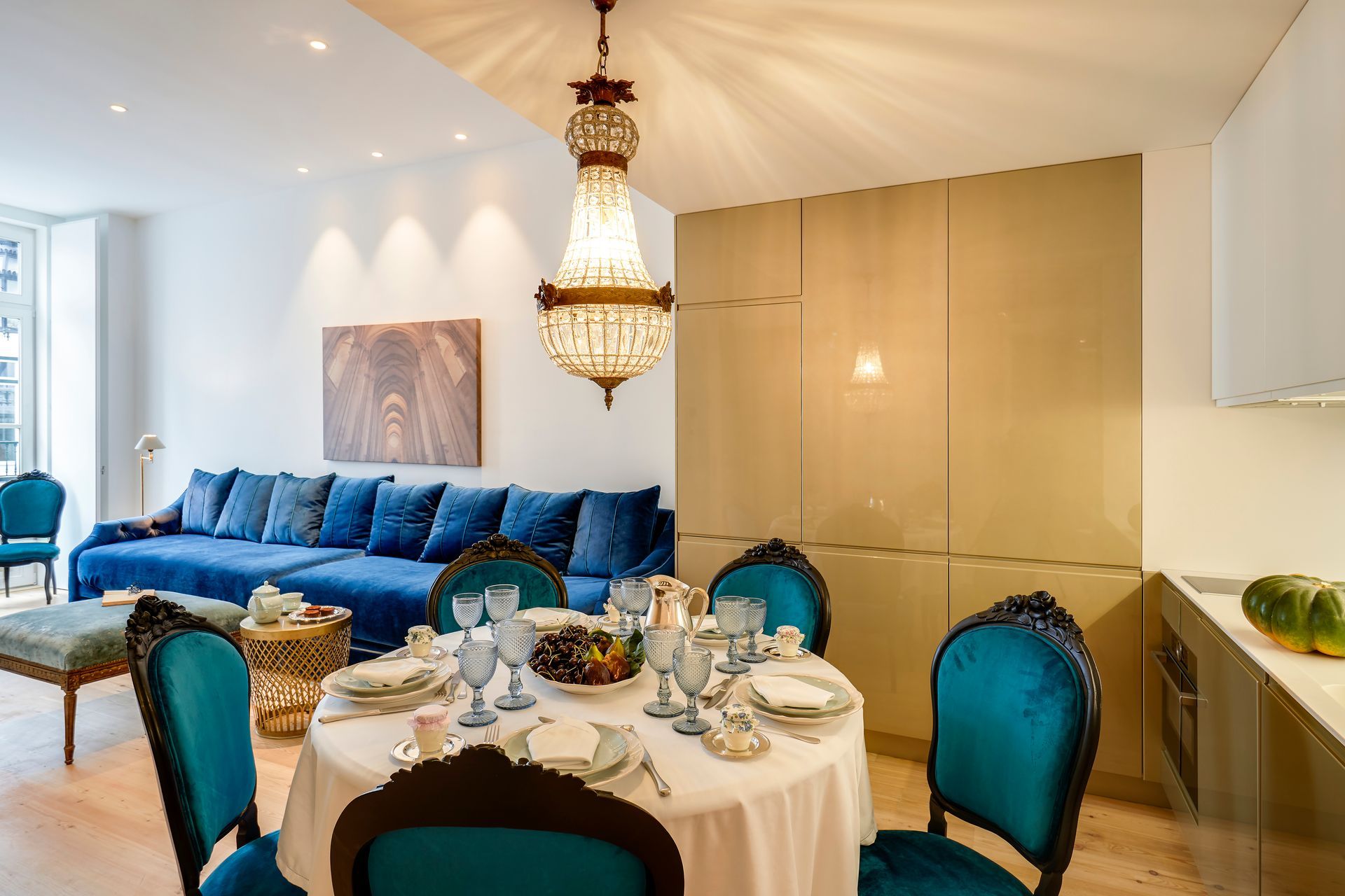 Dining area with round table, blue velvet chairs, blue sofa, ornate chandelier, and light wood flooring.