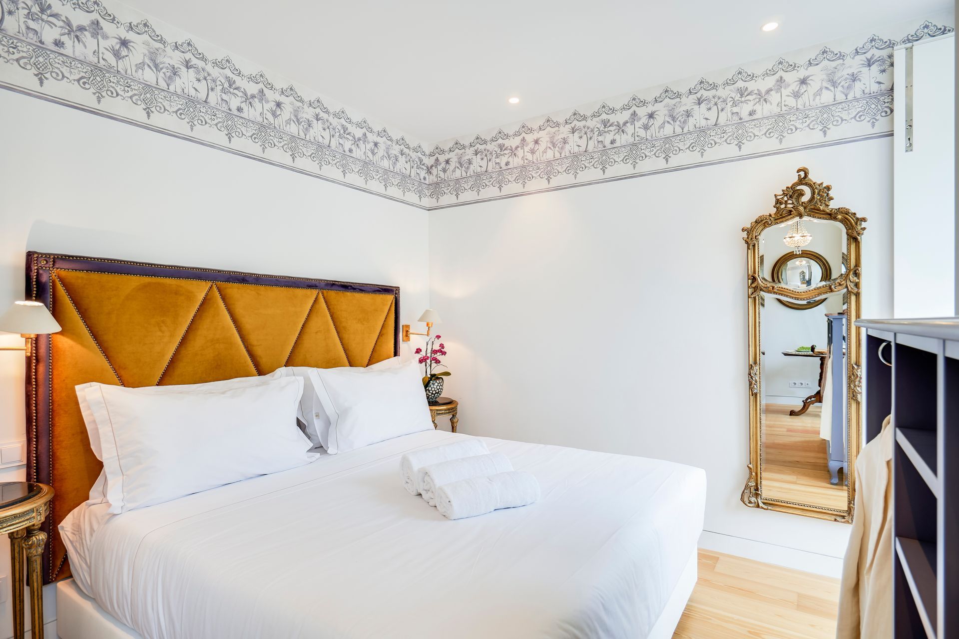 Bedroom with mustard yellow headboard, gold mirror, white bedding, floral wallpaper trim.