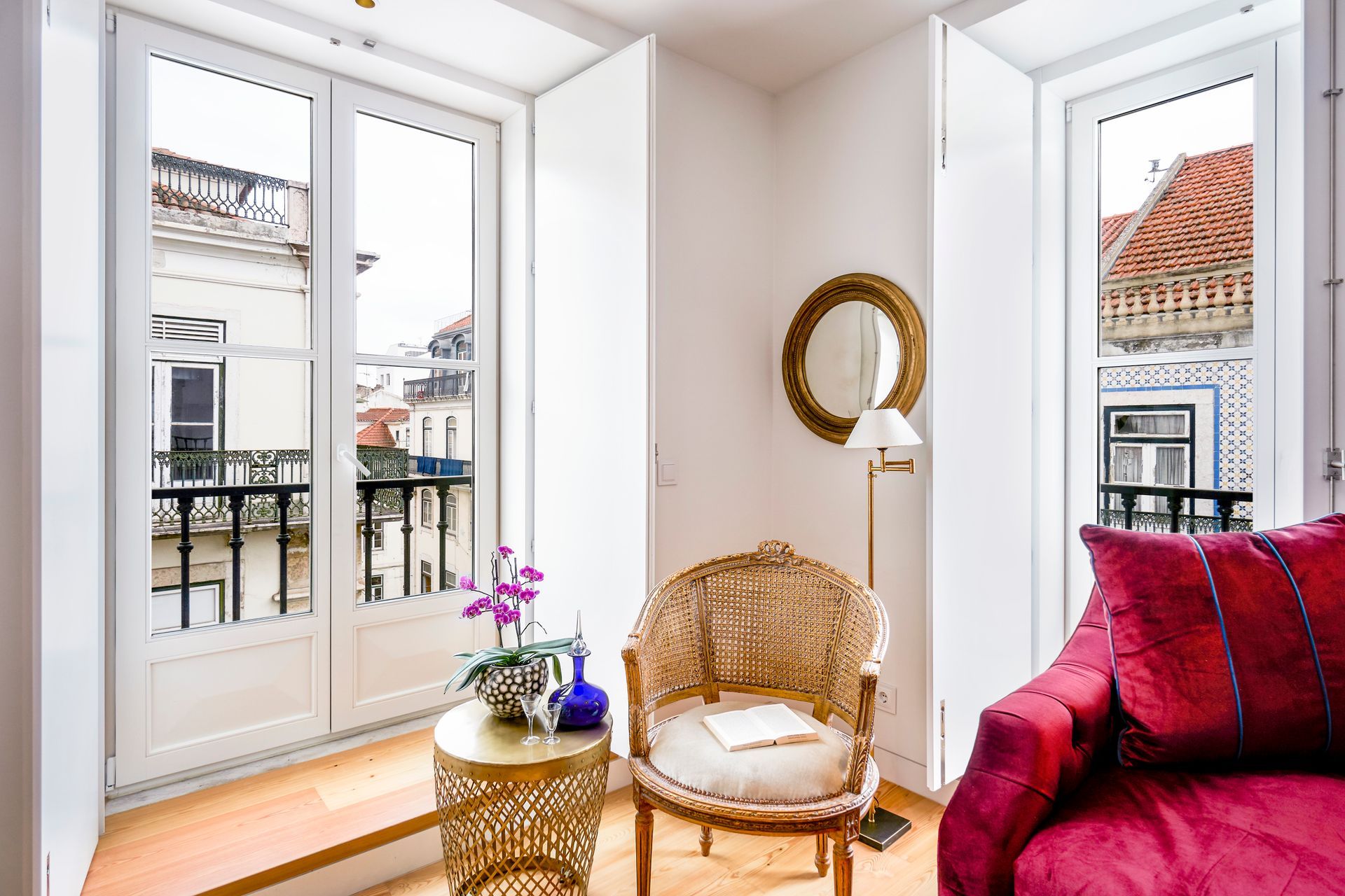 Bright room with balcony view, ornate chair, round mirror, small table, and red sofa.