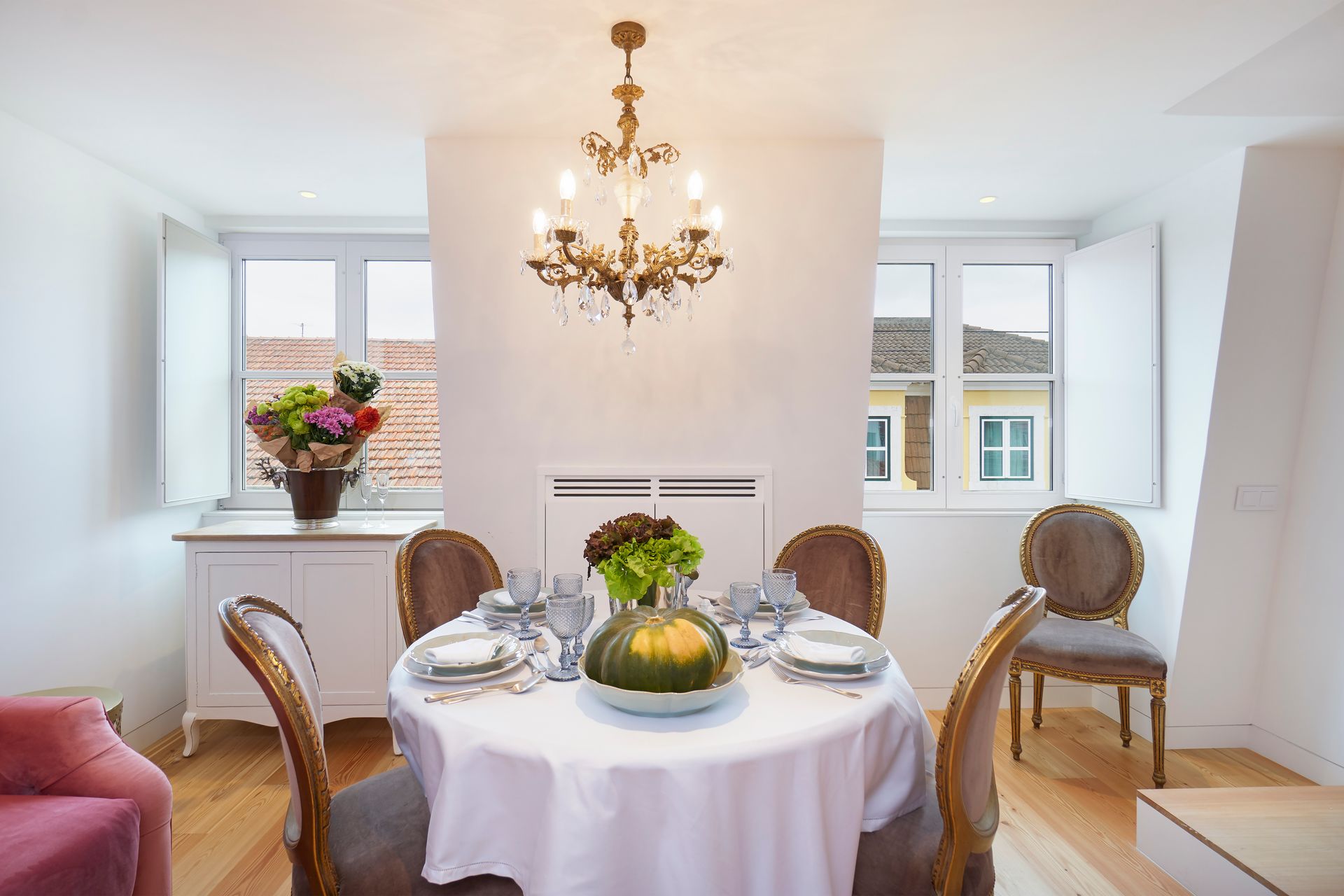 Dining room with round table set for guests, chandelier, windows, and decorative flowers.