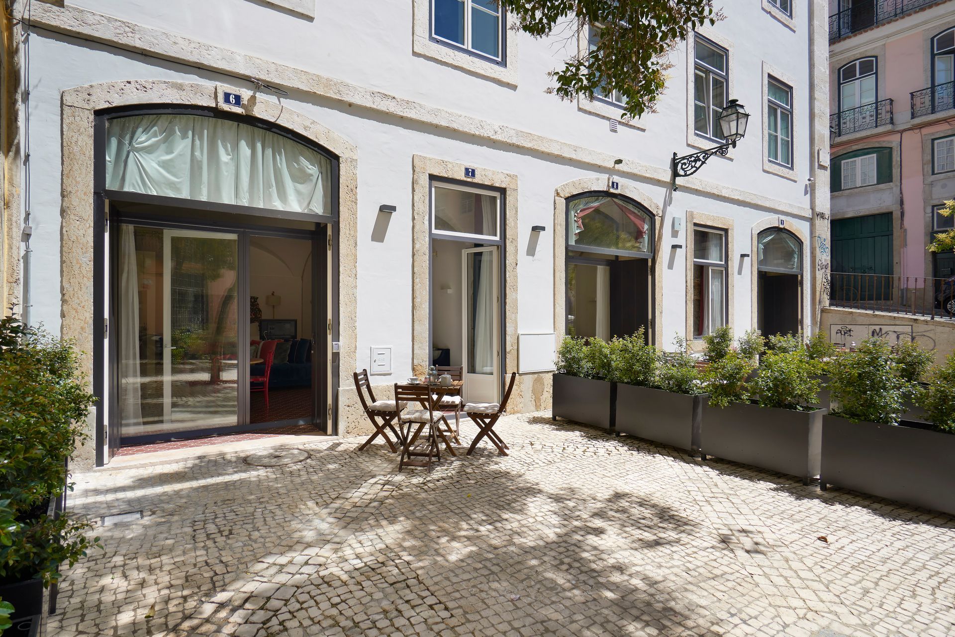 Exterior view of a white building with arched windows, small patio with cafe table and chairs, and potted plants.