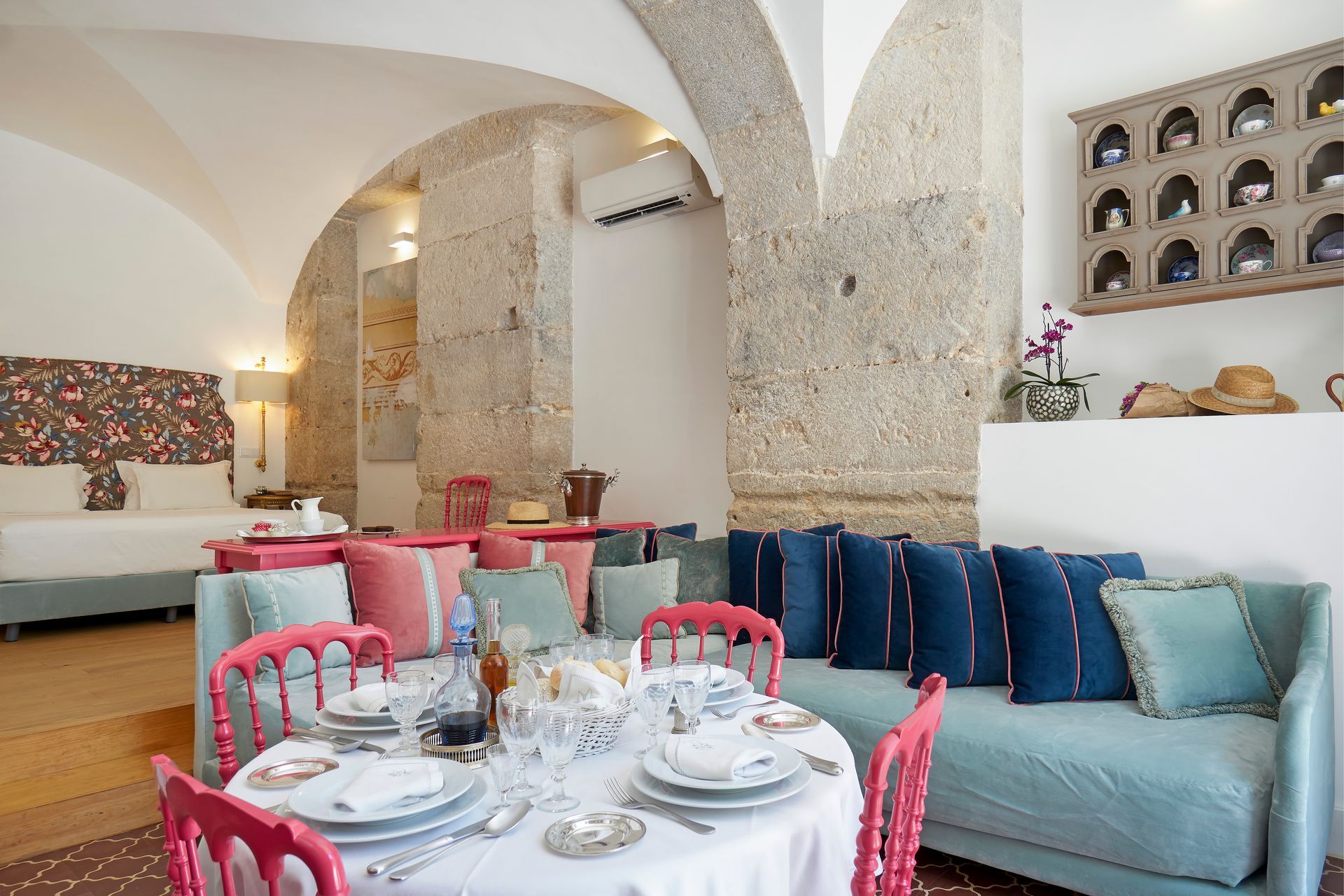 Hotel suite with a dining area and a bed. Light blue sofa with pink chairs, stone pillars.