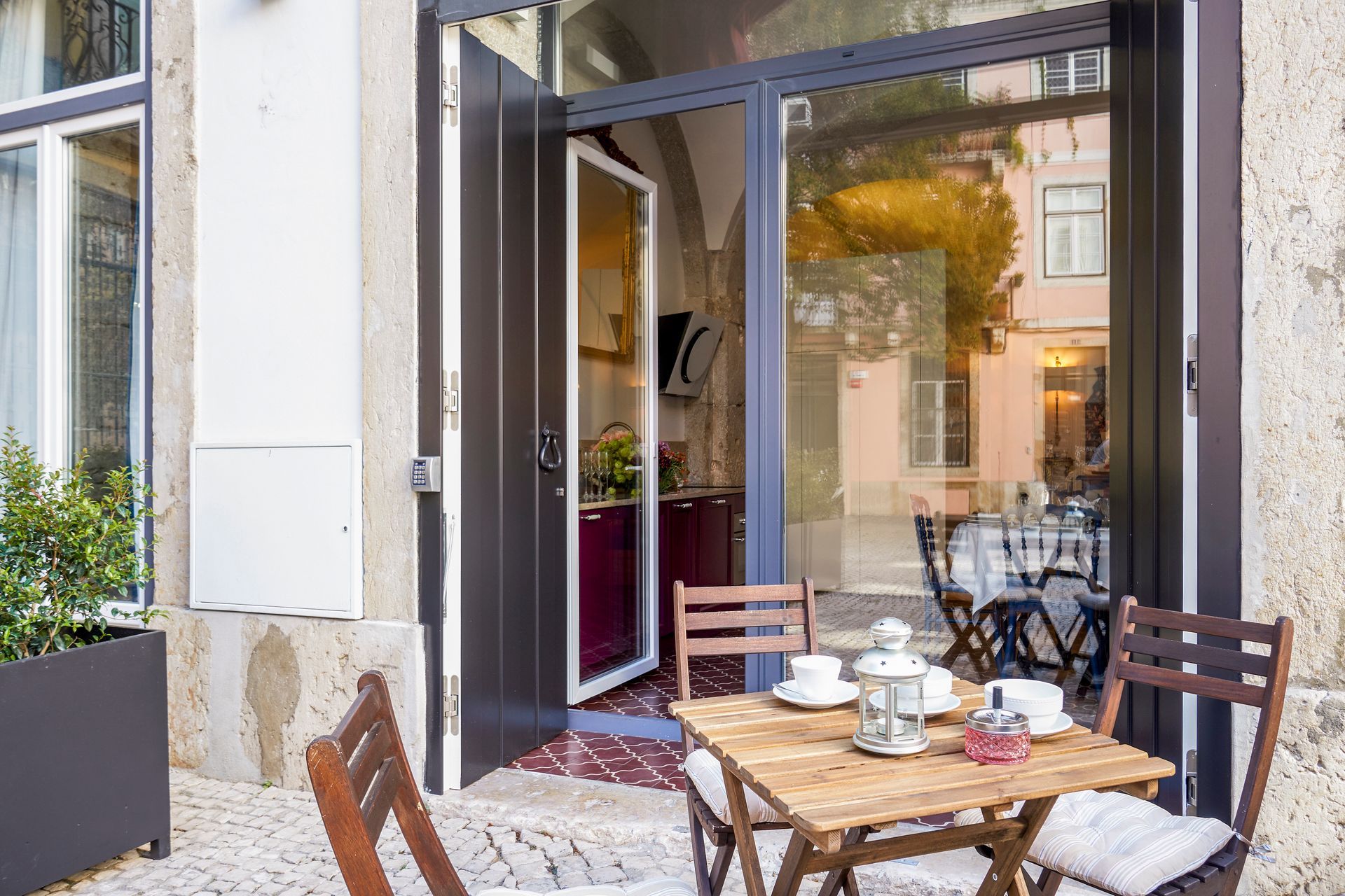 Outdoor cafe scene: table set for tea, doorway to a kitchen visible, cobblestone patio.