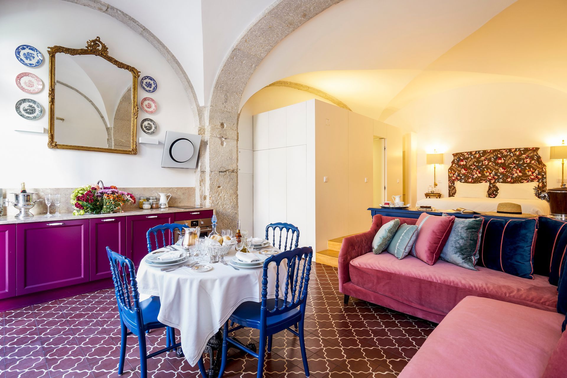 Elegant suite with a dining area, pink sofa, and bed. Purple cabinets, blue chairs, and a large mirror.