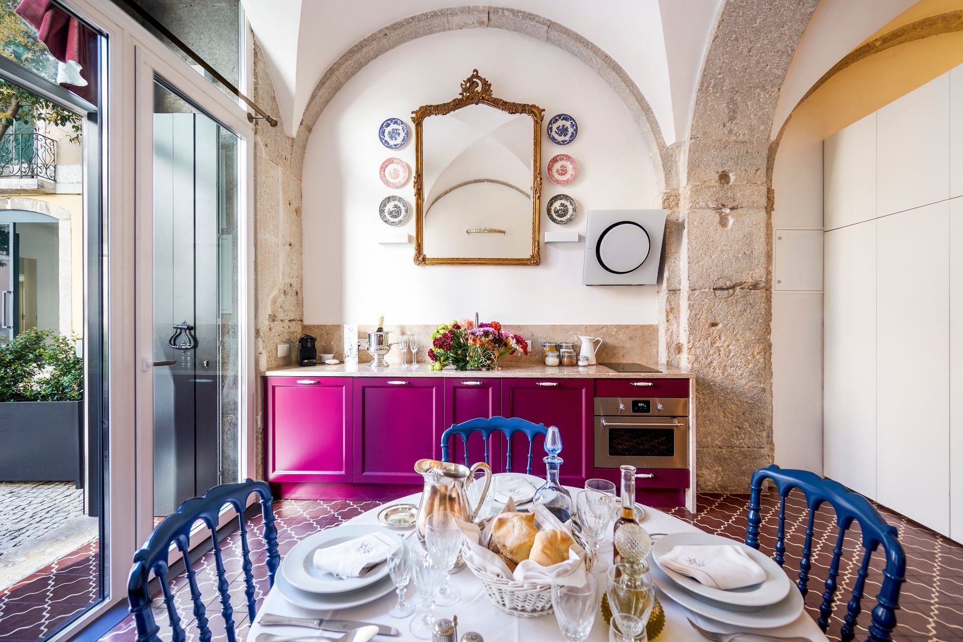 Bright kitchen with fuchsia cabinets, blue chairs, a round table set for a meal, and decorative plates on the wall.