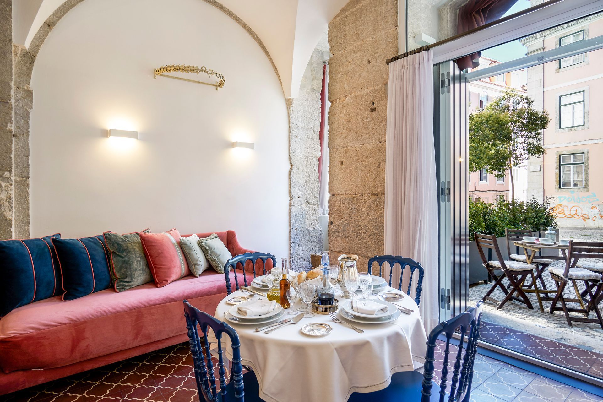 Living room with dining table set for a meal, opening to a patio. Pink sofa, blue chairs, white walls.