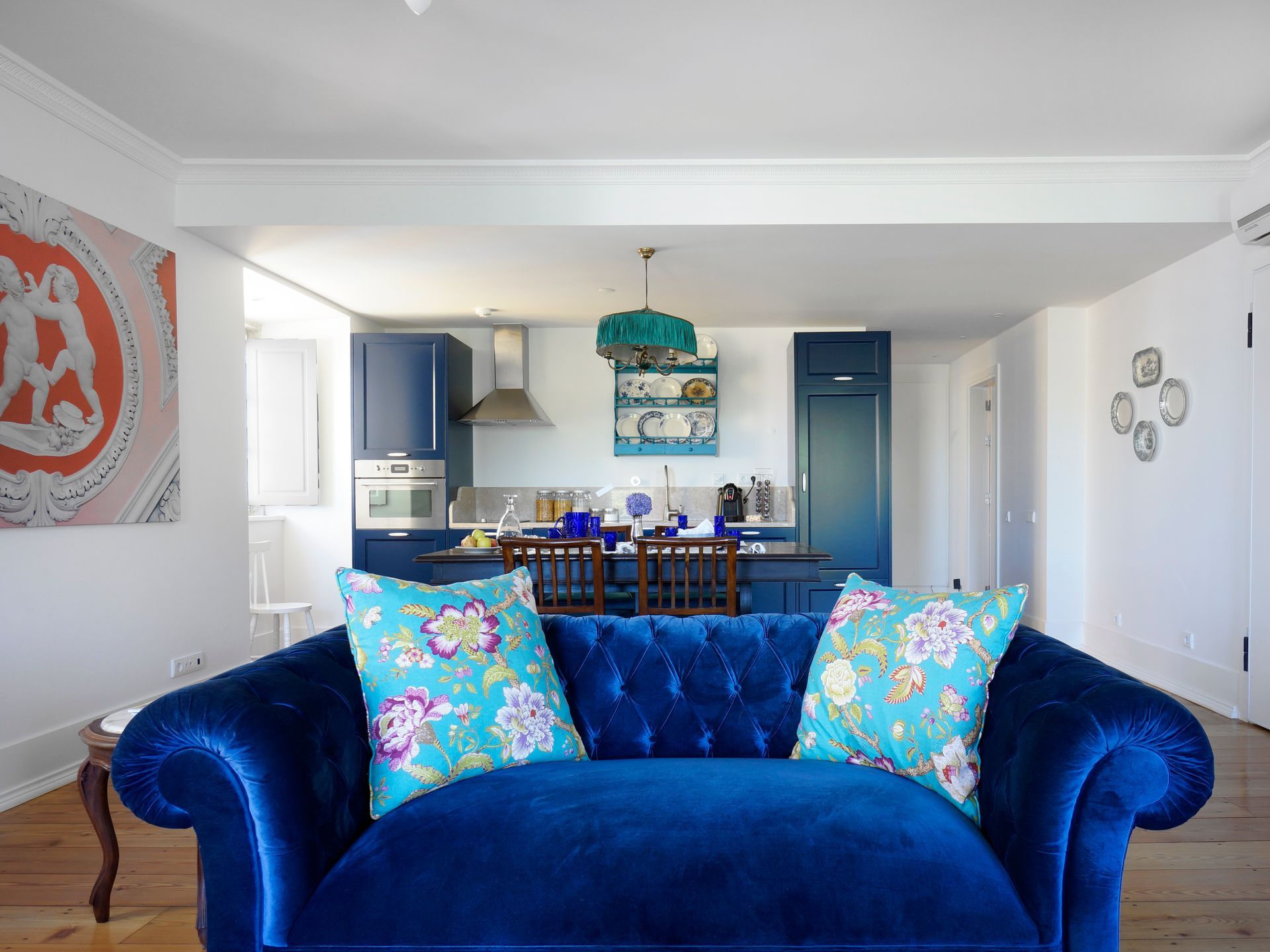 Living area in a premium one-bedroom apartment with blue velvet sofa, colorful cushions, and modern kitchen.