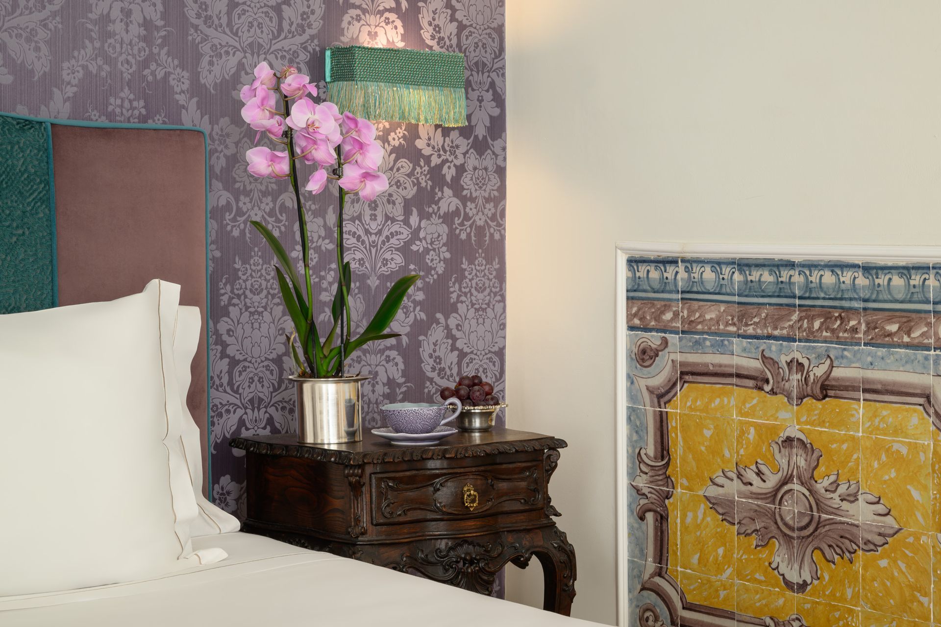 Bedroom scene: Orchid, vintage nightstand, decorative wall panel, purple floral wallpaper, green fringed lamp.