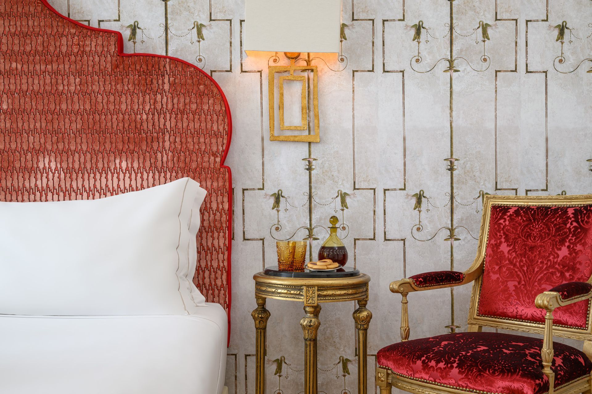 Red wicker headboard and velvet chair in a bedroom with ornate wallpaper, drinks on a side table, and a wall lamp.