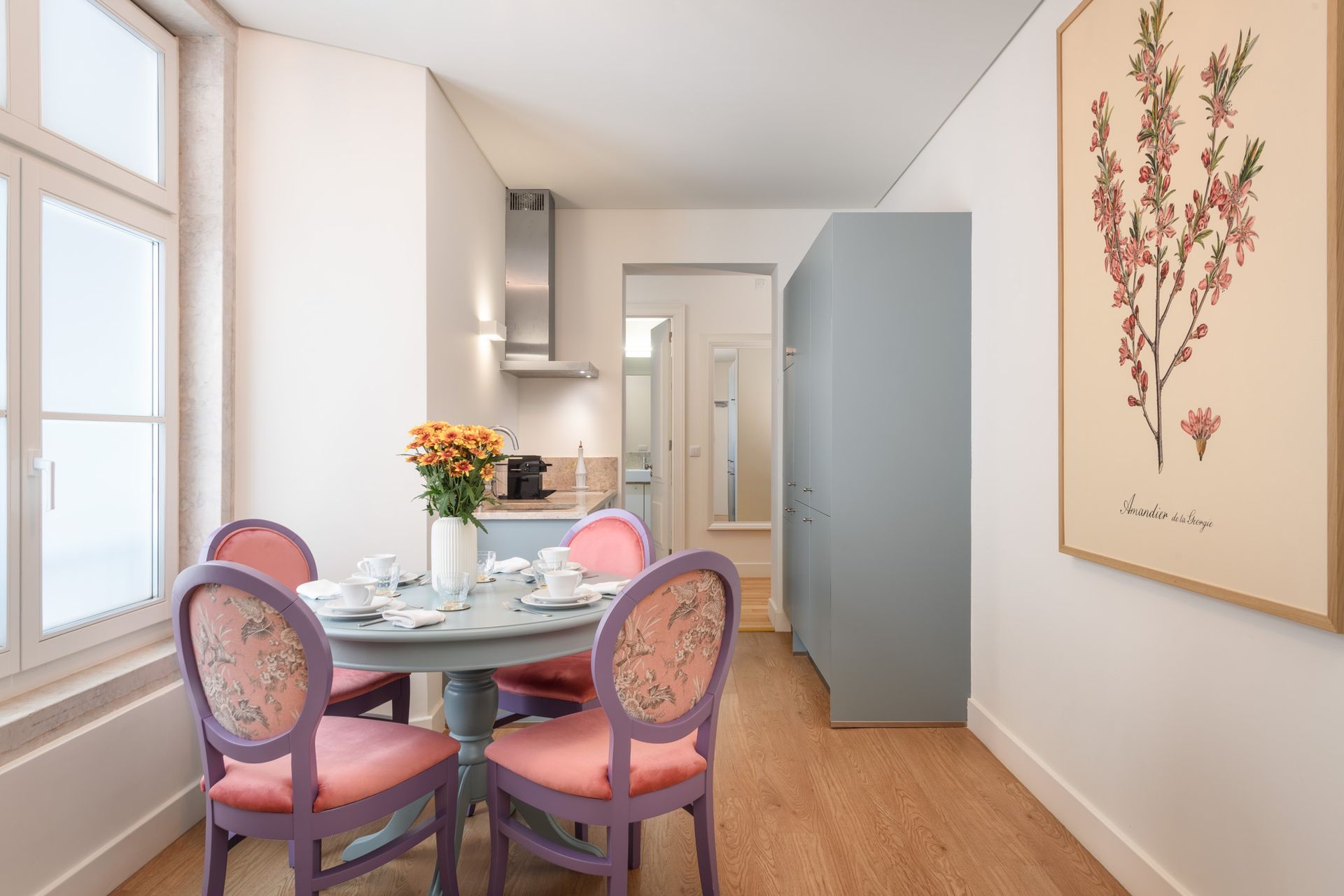 Small dining area with a round table, four pink chairs, and a painting of flowers. Kitchen visible in the background.