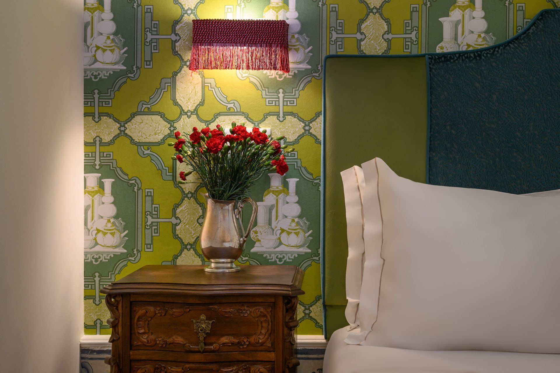 Nightstand with flowers and lamp against patterned wallpaper next to a bed.