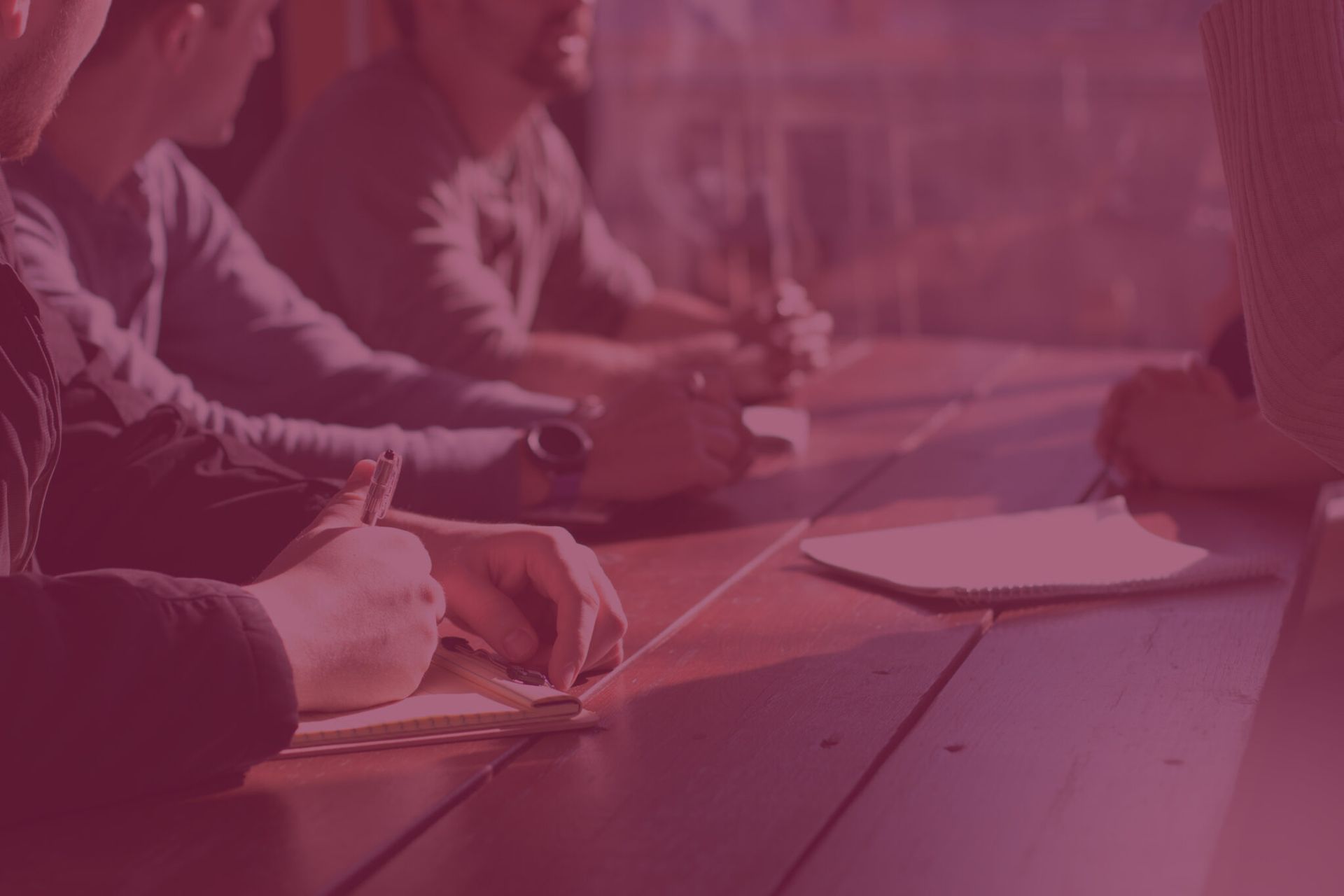 Personas en una reunión, sentadas alrededor de una mesa de madera. Una persona escribe.