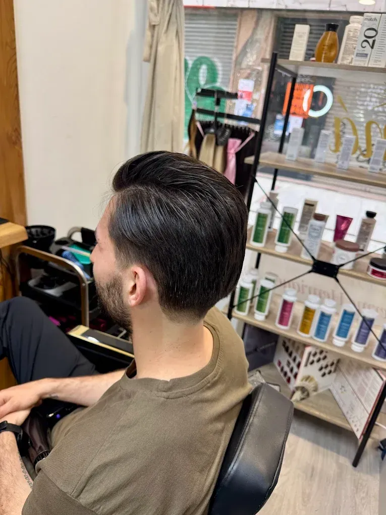 Hombre con cabello oscuro peinado hacia atrás, sentado en una silla de peluquería, visto desde atrás.