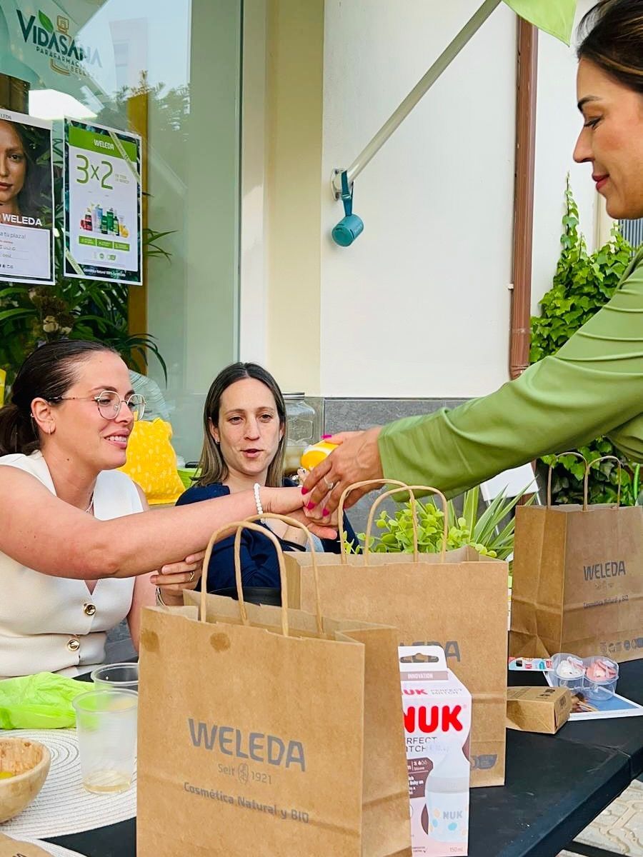 Mujer repartiendo artículos, bolsas de papel marrón, mesa, camisa verde, otras dos mujeres, entorno al aire libre.
