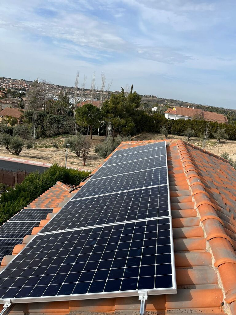Paneles solares instalados sobre un tejado de tejas de terracota, con un paisaje urbano lejano y árboles bajo un cielo nublado.