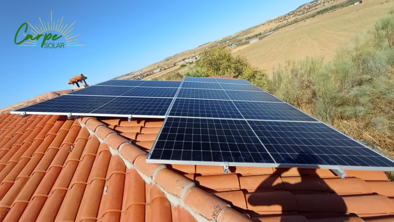 Paneles solares instalados sobre un tejado de tejas rojas, con un logo verde y un fondo de ladera.
