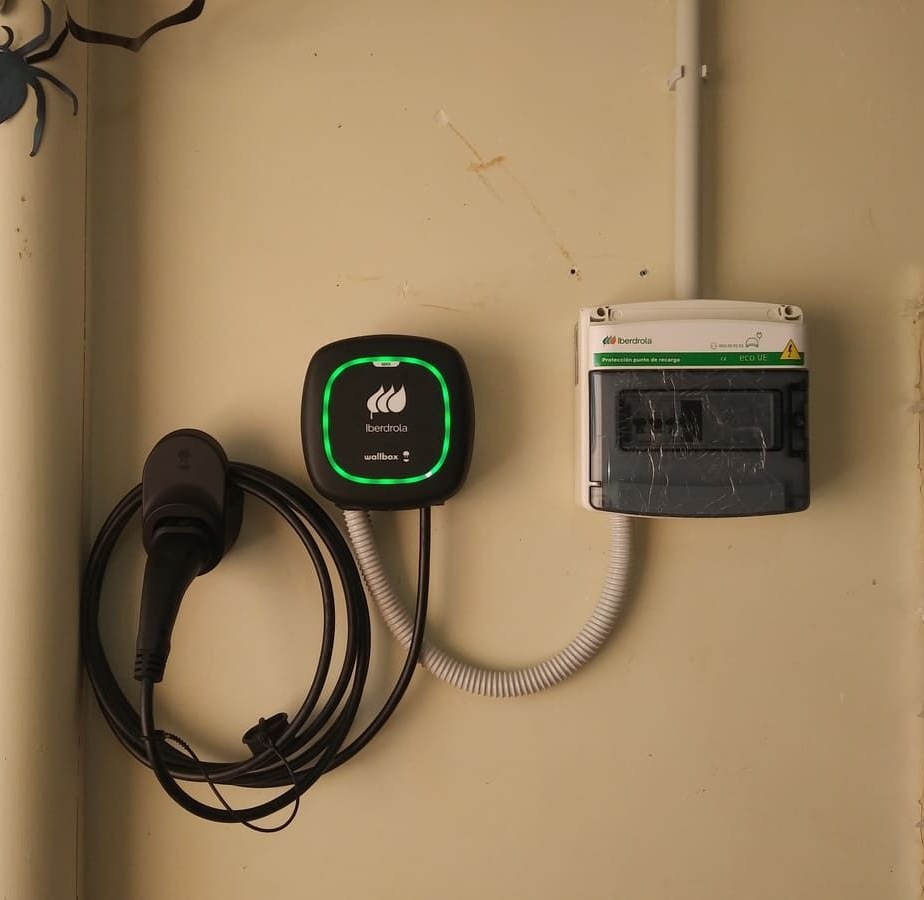 Cargador de vehículo eléctrico montado en la pared con un cable de carga en espiral junto a una caja de interruptores en una pared blanca.