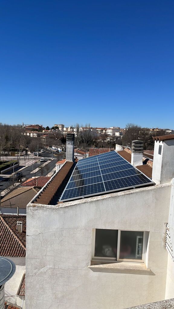 Paneles solares instalados sobre un tejado de tejas rojas contra un cielo azul. También se ven una chimenea y árboles.