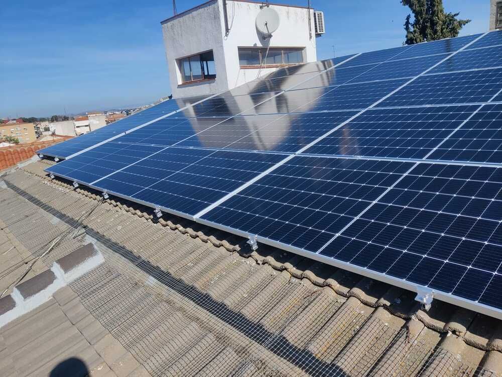 Paneles solares instalados en un tejado de tejas, que reflejan el cielo. Edificio blanco al fondo.