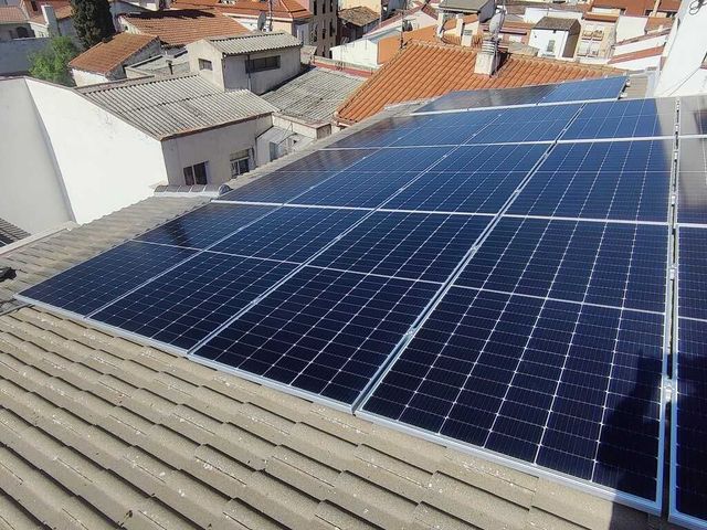 Paneles solares instalados sobre un tejado de tejas rojas contra un cielo azul. También se ven una chimenea y árboles.