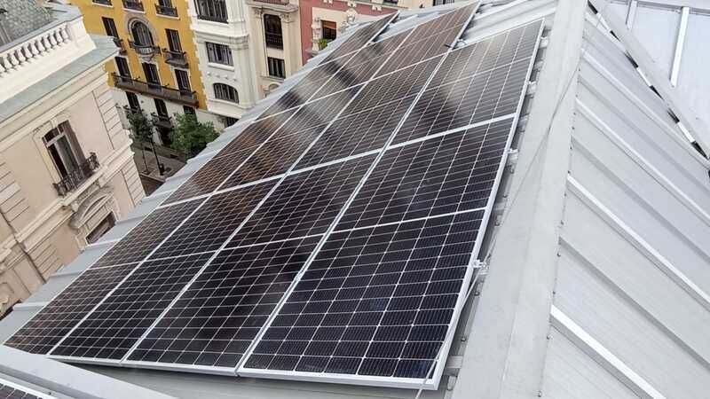 Paneles solares instalados en el techo metálico inclinado de un edificio, que reflejan la luz del sol y tienen como telón de fondo una ciudad.