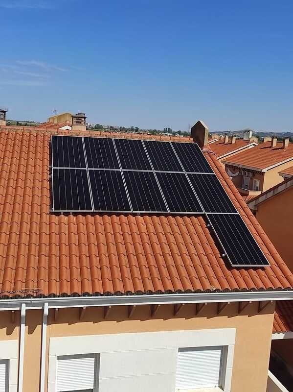 Paneles solares sobre un techo de tejas de terracota contra un cielo azul claro, parte de un edificio en un barrio.