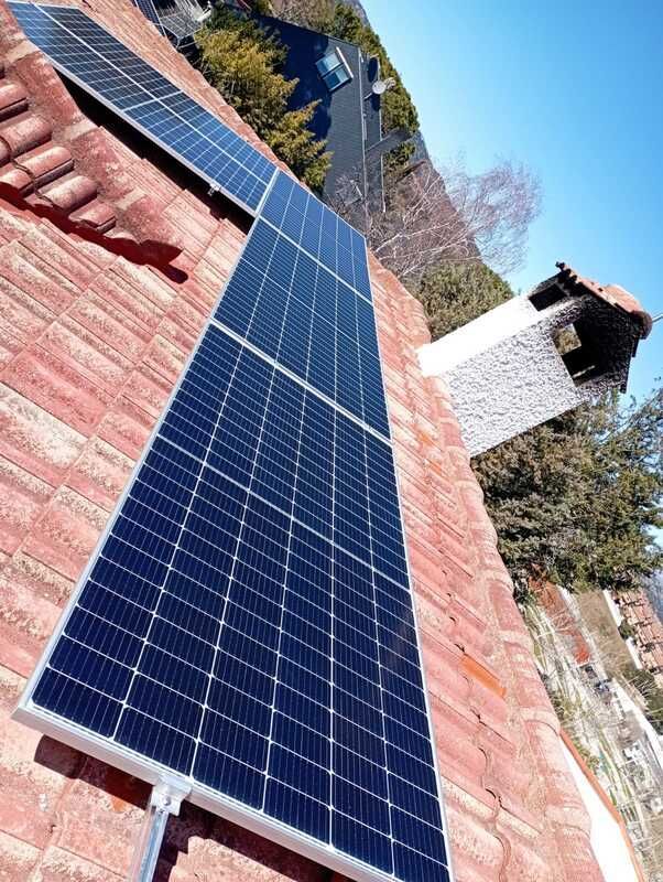 Paneles solares instalados sobre un tejado de tejas rojas contra un cielo azul. También se ven una chimenea y árboles.