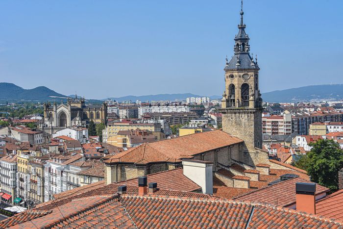 Vista panor谩mica de una ciudad hist贸rica con tejados de tejas rojas y una alta torre de iglesia bajo un cielo azul.