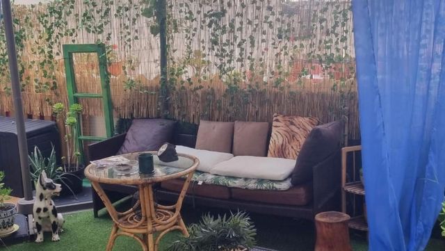 Acogedora zona de estar en el patio con sof谩 de mimbre, mesa de cristal, plantas en macetas y cortina azul.