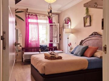 Habitaci贸n luminosa con una cama grande, cortinas rosas y un asiento junto a la ventana en tonos neutros suaves.