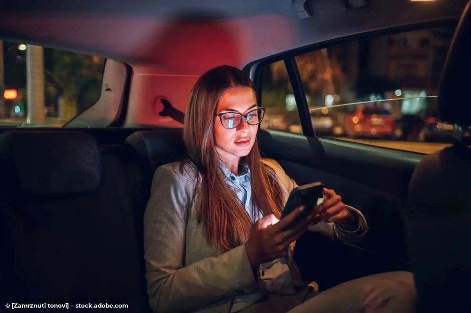 Eine Person mit Brille benutzt nachts auf dem Rücksitz eines Autos ein Smartphone, 