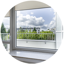 Porte-fenêtre ouverte sur un balcon avec rambarde blanche, donnant sur des arbres verdoyants et un ciel nuageux