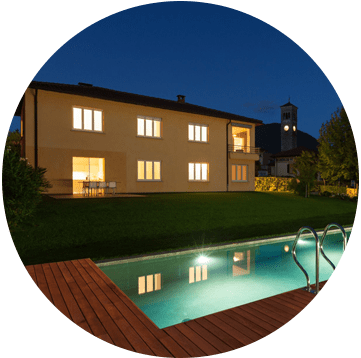 Villa moderne la nuit, au bord d'une piscine éclairée dans le jardin, avec terrasse en bois et reflets.