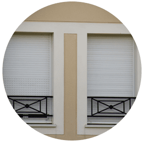 Deux fenêtres blanches aux stores baissés et aux balustrades noires sur une façade beige.