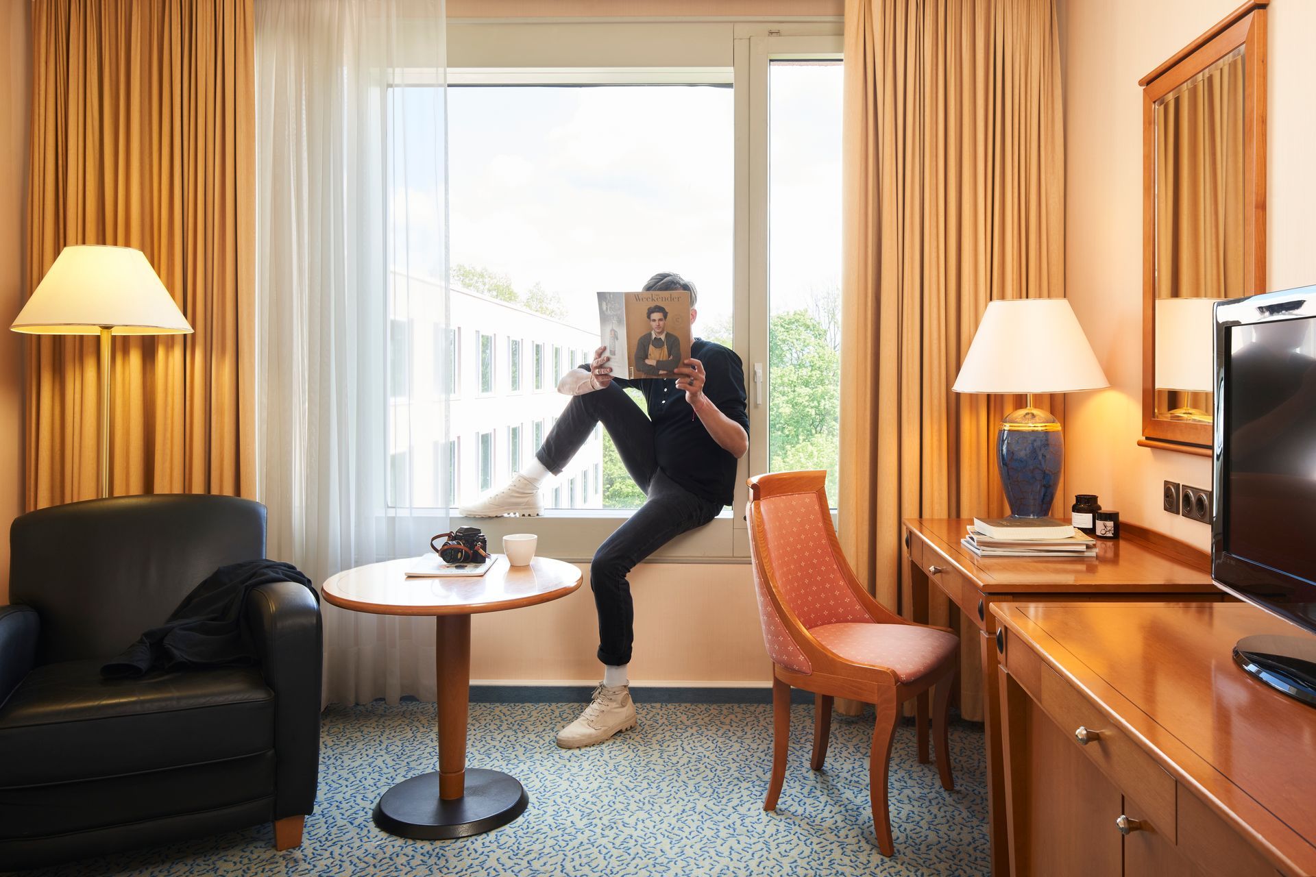 Ein Mann sitzt am Fenster eines Hotelzimmers und liest eine Zeitschrift.