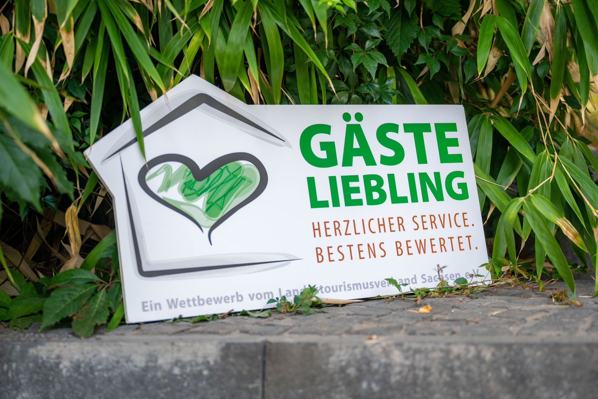 Grüne Werbetafel mit Herzlogo und Text „GÄSTE LIEBLING“ vor dichtem Grün