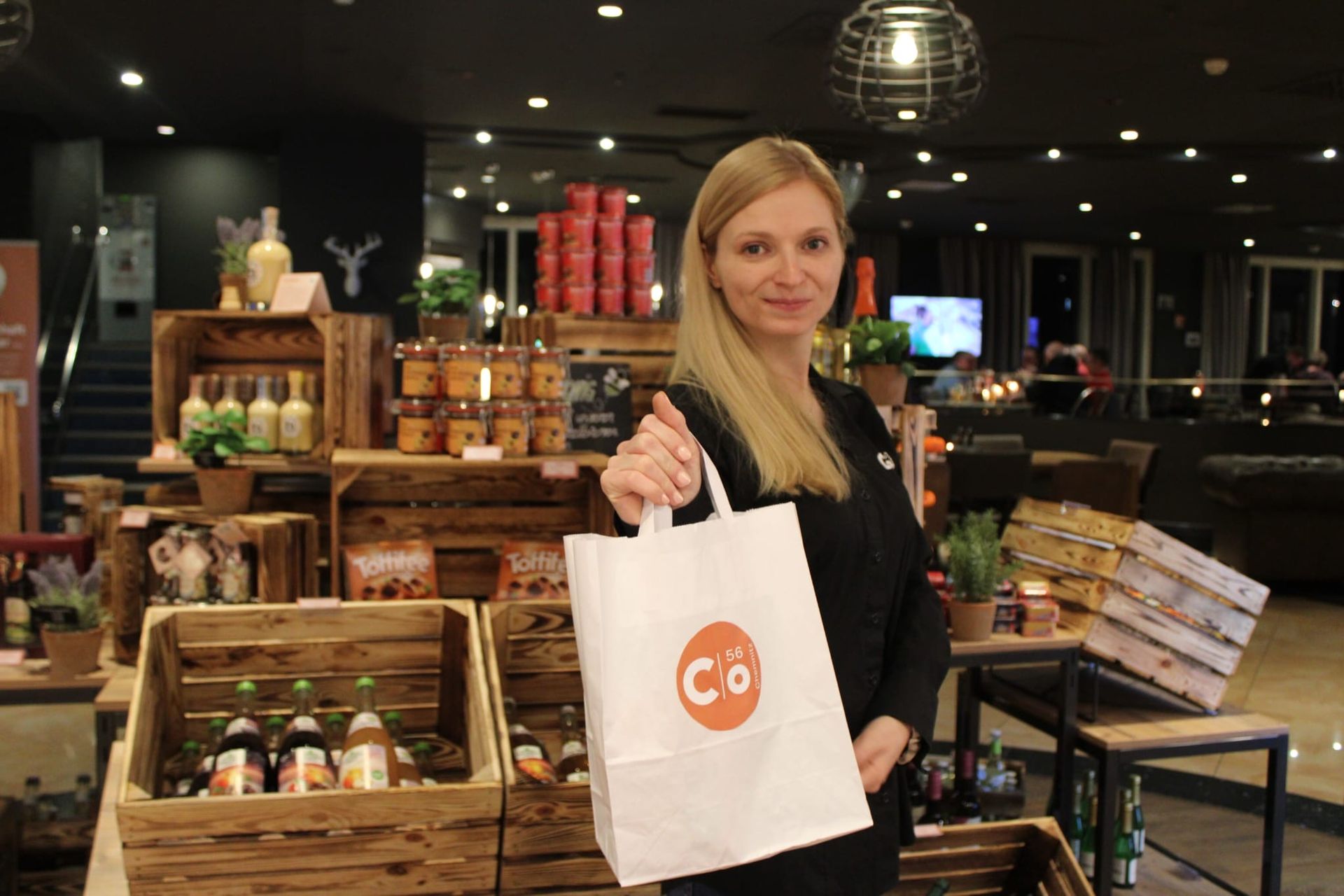 Frau mit einer Einkaufstasche mit Logo in der Hand, steht in der Nähe einer Warenauslage in einem Geschäft.
