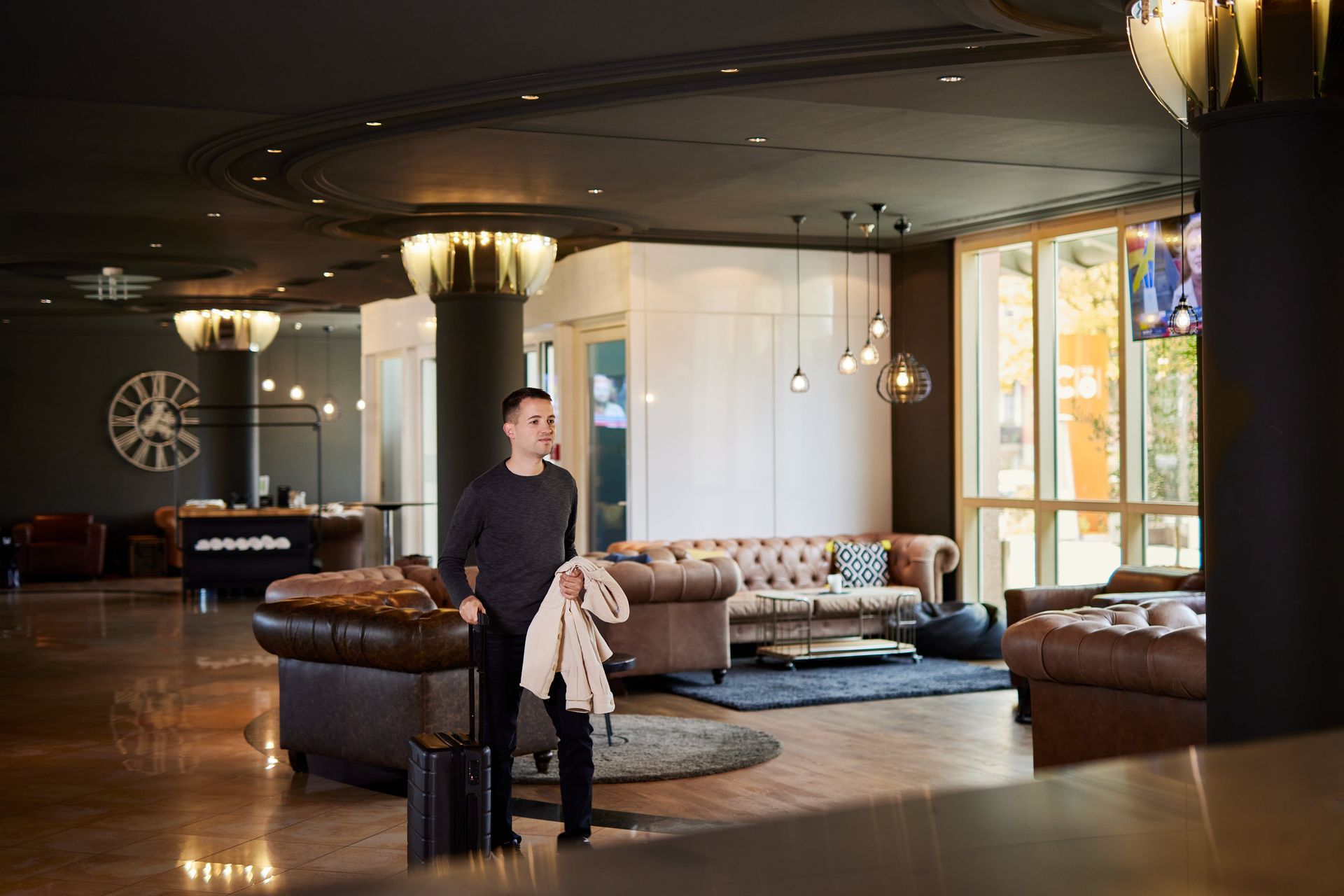 Ein Mann mit Gepäck hält einen Mantel in der Hotellobby. Dunkle Böden, Ledersofas, der Check-in-Schalter ist sichtbar.