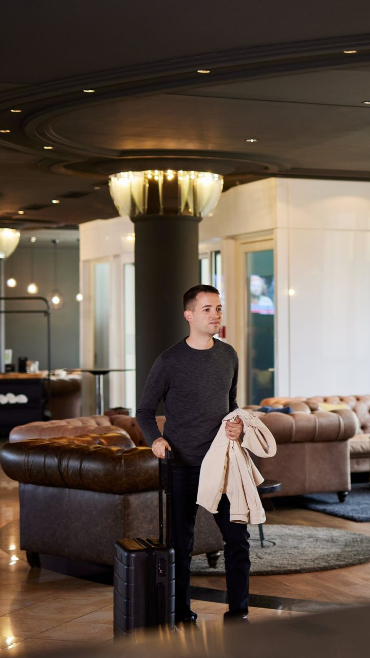 Ein Mann mit Gepäck hält einen Mantel in der Hotellobby. Dunkle Böden, Ledersofas, der Check-in-Schalter ist sichtbar.