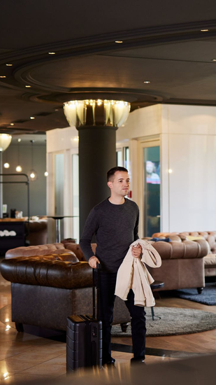 Ein Mann mit Gepäck hält einen Mantel in der Hotellobby. Dunkle Böden, Ledersofas, der Check-in-Schalter ist sichtbar.
