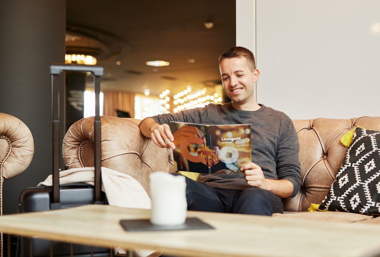 Ein Mann sitzt auf einem braunen Sofa und hält ein Album in der Hand; neben ihm steht Gepäck.
