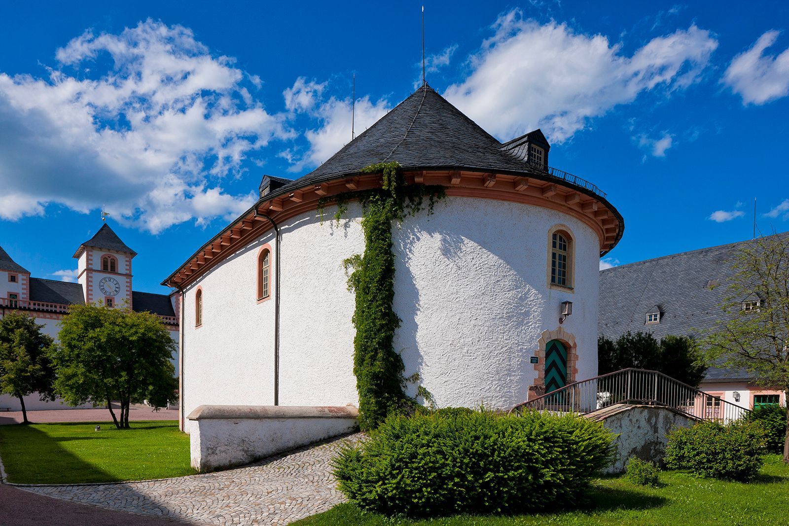 Weißes, rundes Gebäude mit schwarzem Dach, grüner Tür und Kletterpflanzen; sonniger Tag.