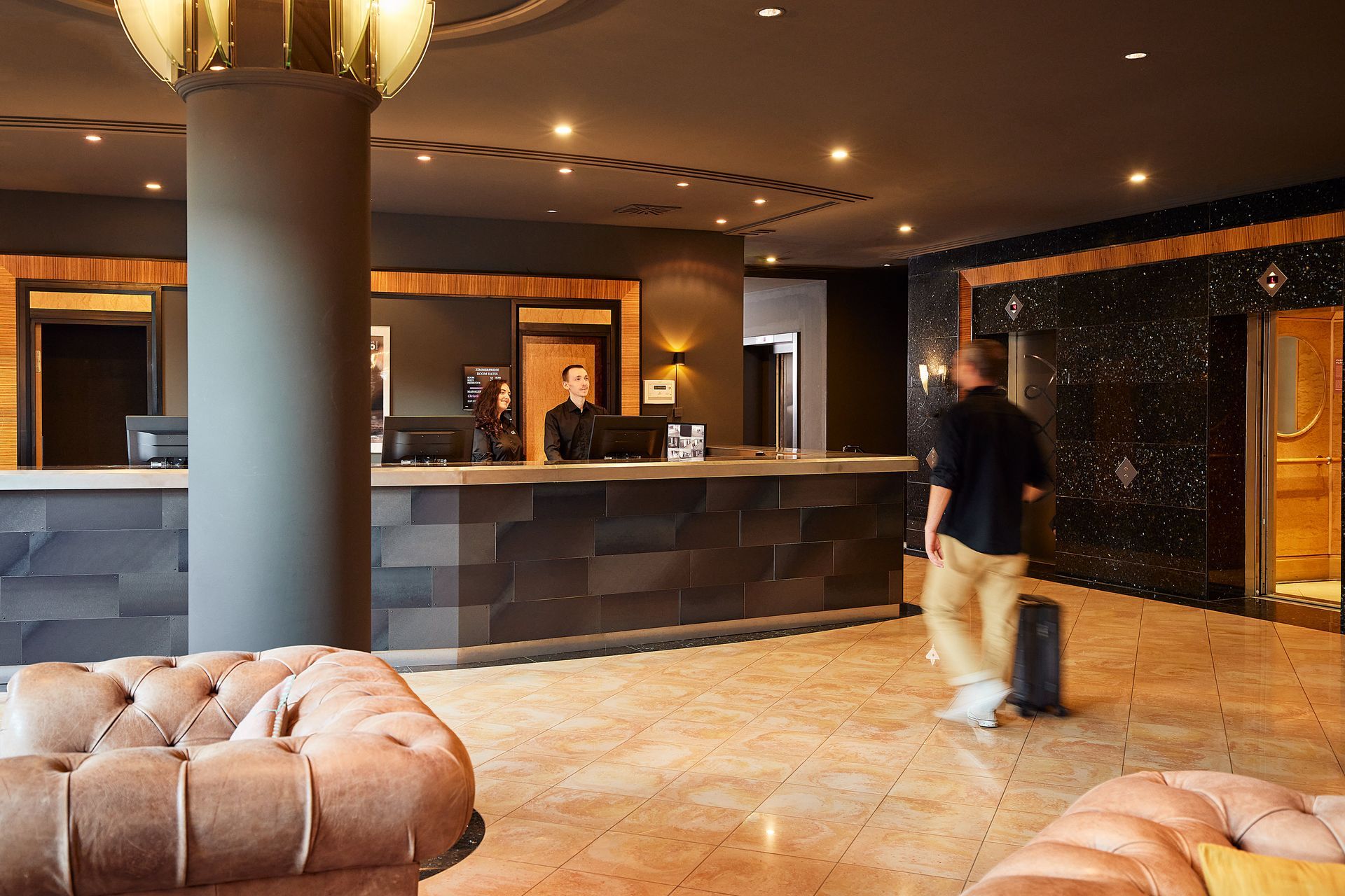 Hotellobby mit Rezeption, einer Person mit Gepäck und Sitzgelegenheiten.